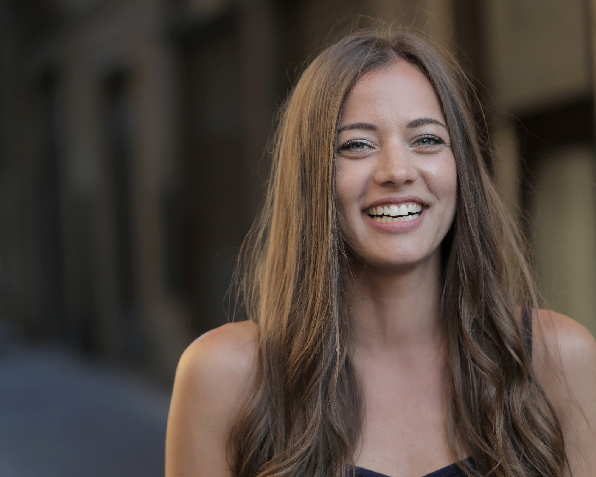 A smiling young woman is sitting on a concrete wall, talking on her mobile phone while smiling at the camera.